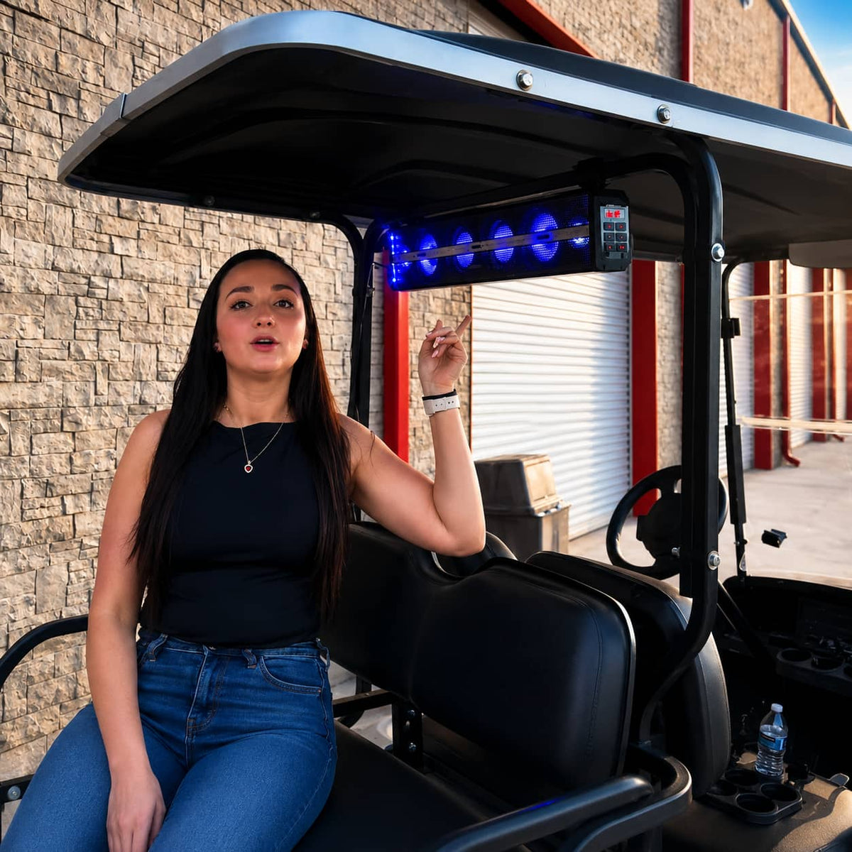 Woman sitting in a custom golf cart pointing at a newly installed roof-mounted Bluetooth golf cart soundbar featuring bright blue LED lights.
